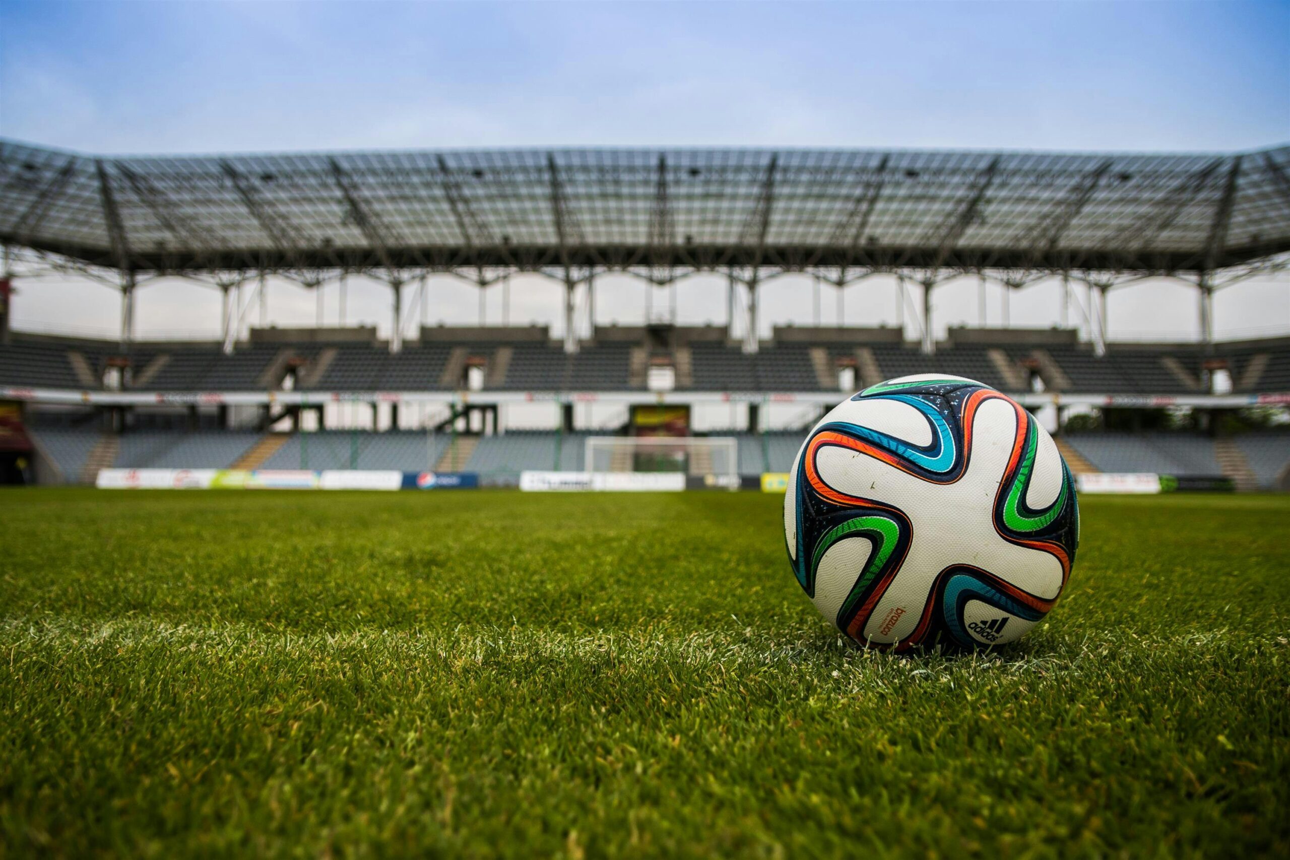 the-ball-stadion-football-the-pitch-46798-46798 Close-up of a soccer ball on a lush grass field with an empty stadium in the background.